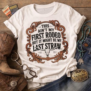 May include: White t-shirt with a brown and tan graphic that reads "This Ain't My First Rodeo But It Might Be My Last Straw." The design includes a steer skull and decorative scrollwork. The shirt is displayed with cowboy boots, a belt buckle, and jewelry.