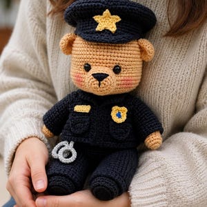Könnte beinhalten: Ein gehäkelter Teddybär in einer dunkelblauen Polizeiuniform, bestehend aus einer passenden Mütze mit gelbem Stern, einer Jacke mit gelbem Abzeichen und einem kleinen Paar hellgrauer Handschellen an der Taille. Der Bär hat ein hellbraunes Fell.