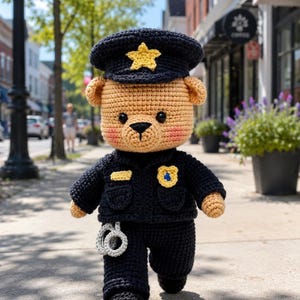 Könnte beinhalten: Ein gehäkelter Teddybär als Polizist verkleidet, mit beigem Körper und rosa Wangen, einer dunkelblauen Uniform, einer passenden Mütze mit gelbem Stern, einem gelben Abzeichen und einem Paar kleiner grauer gehäkelter Handschellen am Gürtel.