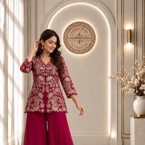 May include: A magenta tunic featuring intricate gold and multi-colored floral embroidery with a scalloped hem and three-quarter sleeves, paired with matching wide-leg trousers. The tunic has a V-neckline and is worn with clear heeled sandals.