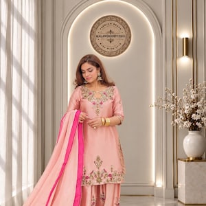 Puede incluir: Un conjunto de salwar kameez rosa claro que presenta una túnica con escote tipo cerradura, mangas tres cuartos y bordados florales intrincados en hilos dorados, verdes y rosas. Los pantalones holgados a juego y el dupatta transparente tienen bordes rosas.