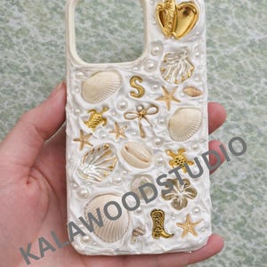 Può includere: Custodia per telefono bianca decorata con conchiglie, perle e dettagli dorati. Presenta un cuore, stelle marine, tartarughe e uno stivale da cowboy. Il testo "KALAWOODSTUDIO" è visibile.