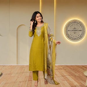 May include: A mustard yellow straight-cut kurta with long sleeves and intricate silver embroidery around the neckline, paired with matching trousers and a sheer, patterned dupatta featuring floral motifs in shades of brown and cream.