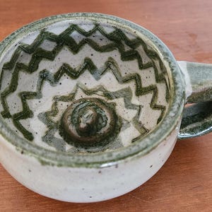 May include: Handmade ceramic shaving mug. The interior has a white base with a green zigzag pattern and a central spiral. The mug has a green handle and rim. A unique design for a luxurious shaving experience.