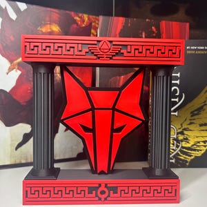 May include: A decorative display piece featuring a red geometric wolf head emblem suspended between two black fluted columns. The top and bottom horizontal bars are red with black Greek key patterns and a central triangular symbol on the top bar.