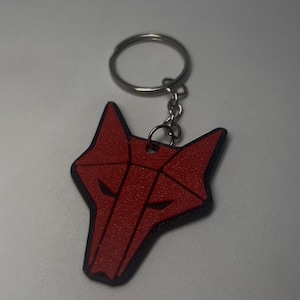 May include: A red wolf head keychain with a black outline. The wolf head is geometric in design and attached to a silver keyring. The keychain is a decorative accessory.