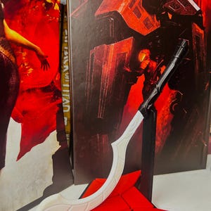 Könnte beinhalten: Ein weiß-schwarzes dekoratives Schwert-Requisit mit roter Basis. Das Schwert hat eine gebogene Klinge und einen schwarzen Griff. Die Basis hat eine rote Sternform. Der Hintergrund zeigt rote und schwarze Kunstwerke.