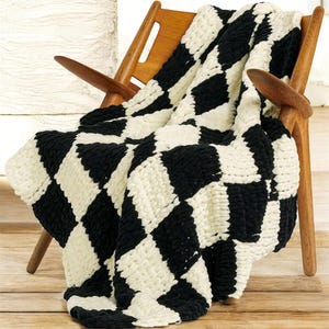May include: A black and cream-coloured, chunky knit blanket with a large checkerboard pattern is draped over a wooden chair. The blanket's thick yarn creates a cosy, textured appearance. The chair has a mid-century modern design.