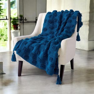 May include: A teal blue knitted throw blanket with a textured, raised pattern and tassels at each corner. The blanket is draped over a cream-colored armchair with dark brown legs, creating a cozy and inviting aesthetic.