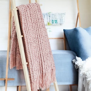 May include: A blush pink knitted throw blanket with fringe, draped over a wooden ladder. The blanket has a textured, ribbed design. A blue sofa and pillows are in the background, creating a cozy, inviting scene. The throw is a home decor item.