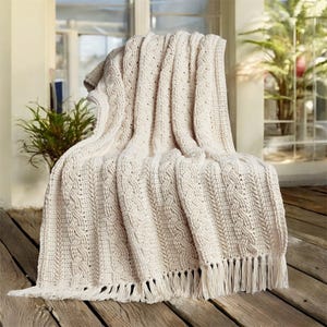 Op de afbeelding: Crèmekleurige gebreide sprei met een textuur kabelbreipatroon en franje. De deken is over een stoel op een houten terras gedrapeerd, waardoor een gezellige en uitnodigende uitstraling ontstaat. De neutrale kleur past bij de natuurlijke buitenomgeving.