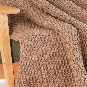 May include: A close-up of a brown knitted throw blanket draped over a wooden chair. The blanket has a textured, ribbed pattern and a warm, neutral color. The chair has a wooden frame and a green cushion.