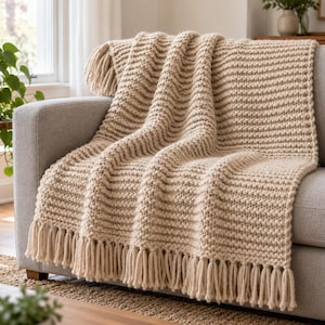 May include: A beige knitted throw blanket with a textured ribbed pattern and long fringe detailing along the edges. The blanket is draped over a gray sofa, showcasing its thick yarn construction and consistent neutral color throughout.