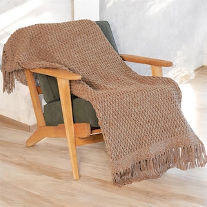 May include: A brown knitted throw blanket with a textured diamond pattern and fringe detail is draped over a wooden armchair with a green cushion. The blanket's warm color and texture create a cozy, inviting aesthetic.