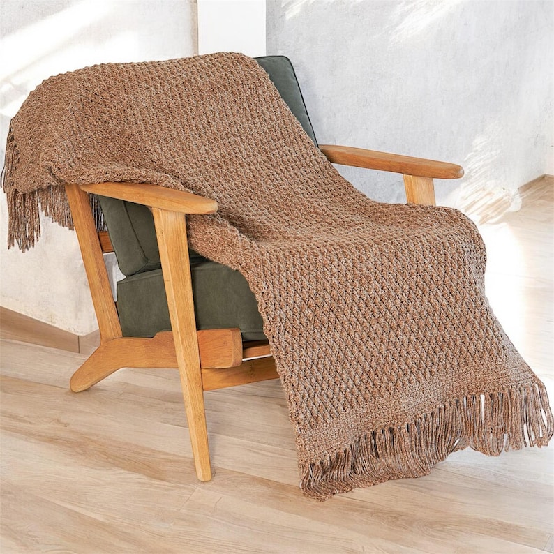 May include: A brown knitted throw blanket with a textured diamond pattern and fringe detail is draped over a wooden armchair with a green cushion. The blanket's warm color and texture create a cozy, inviting aesthetic.