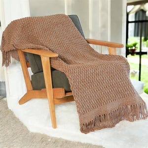 May include: A brown knitted throw blanket with fringe is draped over a wooden armchair with a dark green cushion. The blanket has a textured diamond pattern and is made of a soft, warm material. The chair is set on a white rug.