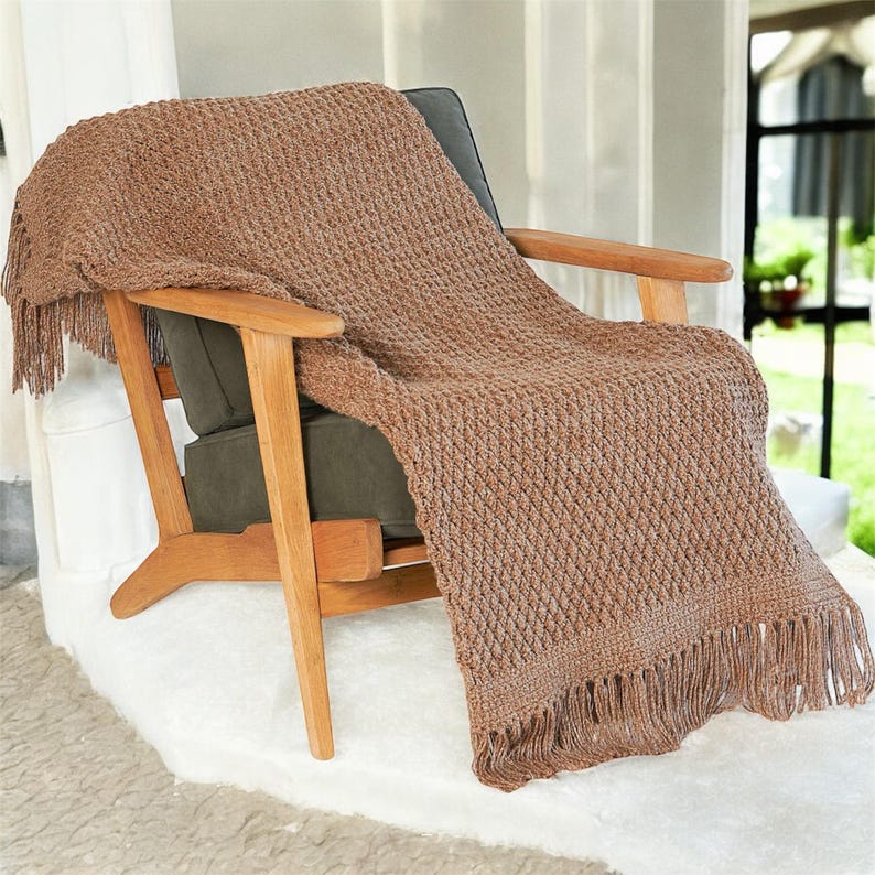 May include: A brown knitted throw blanket with fringe is draped over a wooden armchair with a dark green cushion. The blanket has a textured diamond pattern and is made of a soft, warm material. The chair is set on a white rug.