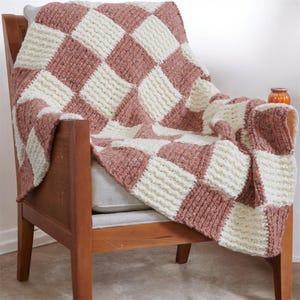 May include: A cozy, handmade blanket draped over a wooden chair. The blanket features a checkerboard pattern of alternating squares in cream and dusty rose. The textured knit adds visual interest and warmth to the home decor.