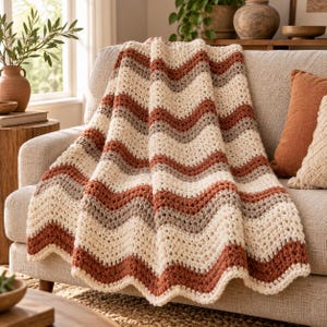 May include: A crocheted throw blanket featuring a repeating chevron pattern in alternating horizontal stripes of cream, terracotta orange, and light taupe. The textile has a thick, textured yarn construction with a wavy, scalloped bottom edge.