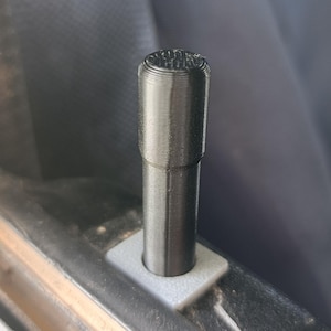 May include: A black, cylindrical car door lock pin with a textured surface, mounted on a square, light gray base. The pin is vertically oriented, and the image is a close-up shot.