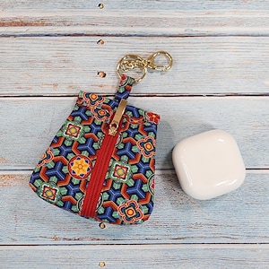 May include: A small, patterned pouch with a gold zipper and keychain next to a white, square wireless earbud case. The pouch features a colorful geometric design with red, blue, yellow, and green accents. The background is a light blue wooden surface.