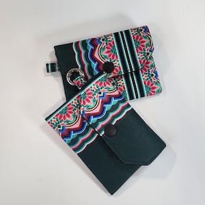May include: Two dark green wallets with a colorful floral pattern. The wallets have a flap closure with a black button and a silver ring for attaching a key chain. The floral pattern includes red, pink, blue, and green.