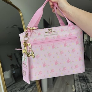May include: A light pink soft cooler bag featuring a floral pattern with small pink roses and yellow accents. The bag displays the Stanley and LoveShackFancy logos in gold text. It includes a pink zip, two top handles, and gold charms.