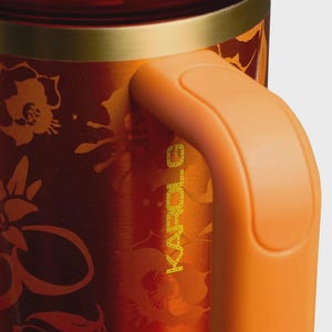 May include: A metallic orange and red tumbler featuring a floral pattern and the text KAROL G printed vertically. The drinkware has a gold-toned rim and a solid orange handle attached to the side, showcasing a smooth, reflective finish throughout.