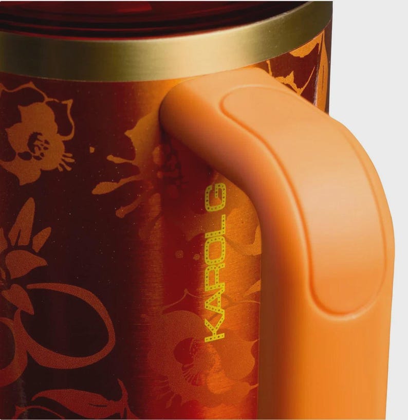 May include: A metallic orange and red tumbler featuring a floral pattern and the text KAROL G printed vertically. The drinkware has a gold-toned rim and a solid orange handle attached to the side, showcasing a smooth, reflective finish throughout.