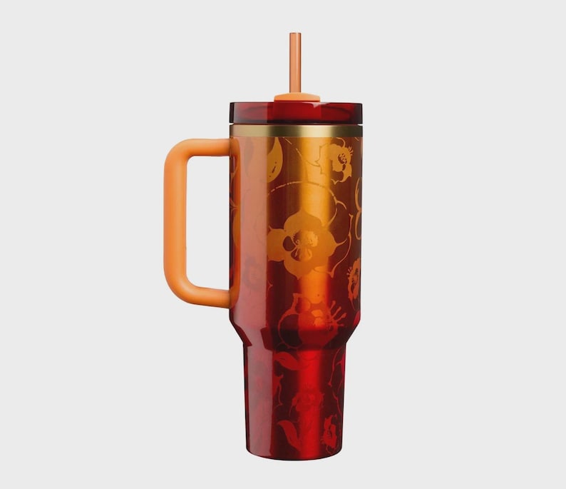 May include: A tall, cylindrical tumbler with a deep red and orange gradient finish featuring a subtle floral pattern. It includes a sturdy orange side handle, a matching orange straw, and a dark red lid with a metallic gold-toned rim accent.