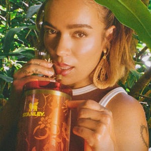 May include: A copper-colored Stanley tumbler featuring a floral pattern and the Stanley logo in yellow. The tumbler has a translucent red lid and a matching straw. The design includes a metallic finish with a repeating botanical motif across the body.