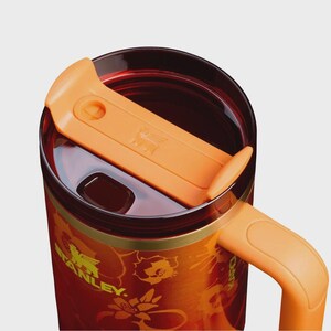 May include: A stainless steel travel mug with a deep red and orange gradient finish featuring a floral pattern and the Stanley logo in yellow. It includes a translucent dark red lid with an orange sliding closure and an orange side handle.