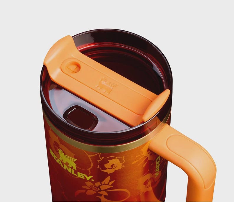May include: A stainless steel travel mug with a deep red and orange gradient finish featuring a floral pattern and the Stanley logo in yellow. It includes a translucent dark red lid with an orange sliding closure and an orange side handle.