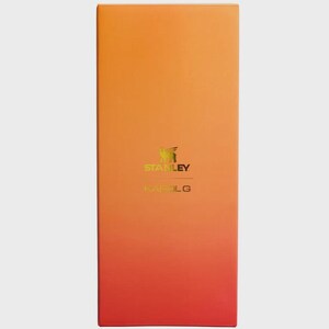 May include: A rectangular product box featuring a vertical gradient that transitions from a bright orange at the top to a deep red at the bottom. The center displays the gold-colored Stanley logo and the text KAROL G printed in a clean font.