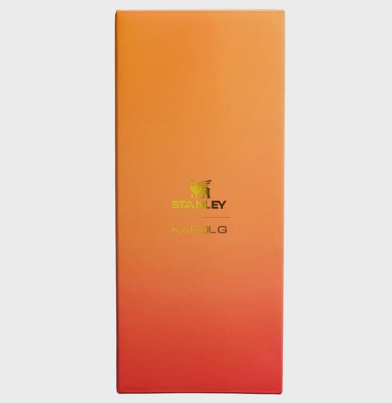 May include: A rectangular product box featuring a vertical gradient that transitions from a bright orange at the top to a deep red at the bottom. The center displays the gold-colored Stanley logo and the text KAROL G printed in a clean font.