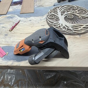 May include: A black and orange power sander with a black sanding pad is on a light brown surface. A carved wooden tree of life design is in the background. A pencil and other tools are also visible.