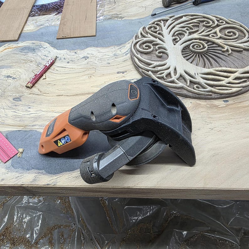 May include: A black and orange power sander with a black sanding pad is on a light brown surface. A carved wooden tree of life design is in the background. A pencil and other tools are also visible.