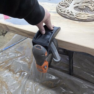 May include: A black and orange power sander is being used on a wooden surface. The sander is held in place by a hand. The wooden surface is light brown and has a natural grain. Sawdust is visible.