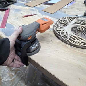 May include: A Ridgid brand orbital sander in gray and orange is held over a wooden surface. A carved wooden tree of life design and other woodworking tools are visible in the background. The sander is being used for a woodworking project.