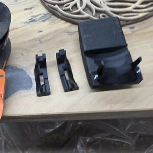 May include: Black plastic components, likely for a tool or machine. The image shows two small brackets and a larger curved piece, all in a matte black finish. Fasteners are visible on the larger piece. The items are on a light-colored wooden surface.