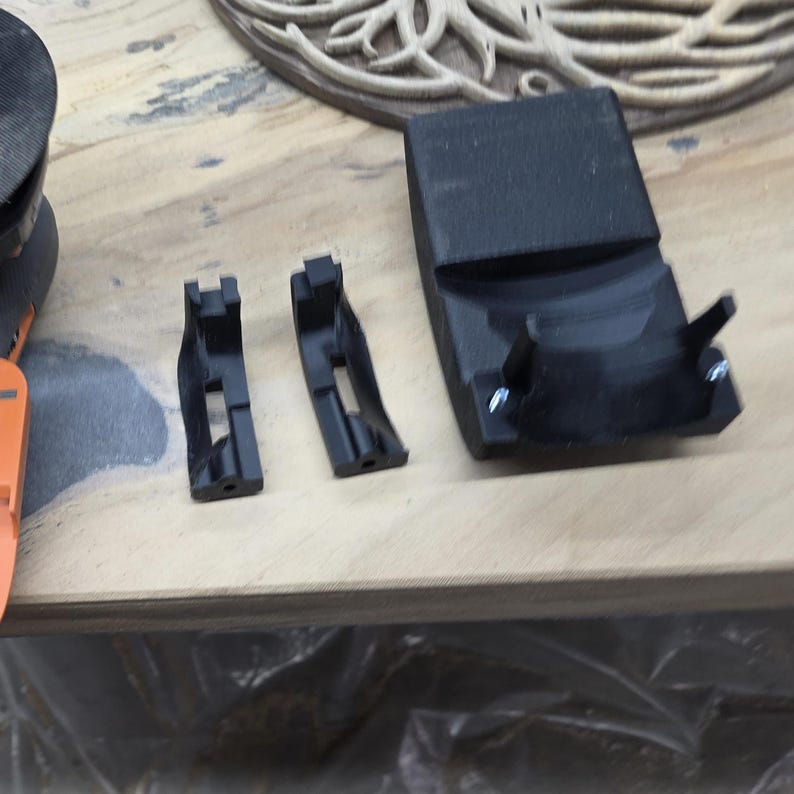 May include: Black plastic components, likely for a tool or machine. The image shows two small brackets and a larger curved piece, all in a matte black finish. Fasteners are visible on the larger piece. The items are on a light-colored wooden surface.