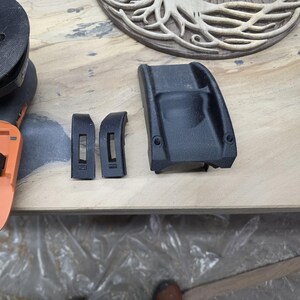 May include: Black plastic components, including two curved pieces with rectangular cutouts and a larger, contoured piece with two circular holes, are displayed on a light-colored wooden surface. A portion of an orange and black tool is visible.