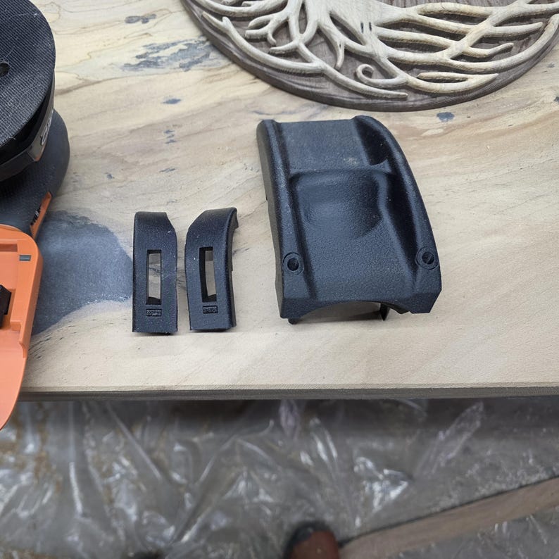 May include: Black plastic components, including two curved pieces with rectangular cutouts and a larger, contoured piece with two circular holes, are displayed on a light-colored wooden surface. A portion of an orange and black tool is visible.