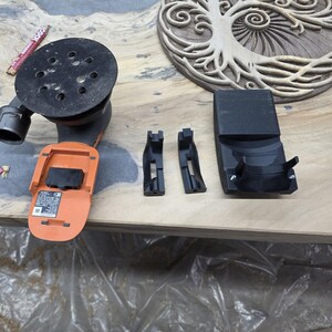 May include: An orange and black power sander with a black sanding pad. Two black plastic brackets and a black charging station are also present. The items are on a wooden surface, with a carved wooden tree of life design in the background.