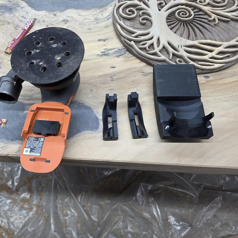 May include: An orange and black power sander with a black sanding pad. Two black plastic brackets and a black charging station are also present. The items are on a wooden surface, with a carved wooden tree of life design in the background.