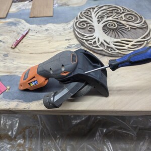 May include: An orange and black power sander with a blue screwdriver. A wooden carving of a tree is in the background. The sander is on a wooden surface.