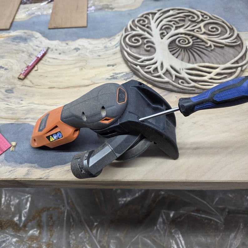 May include: An orange and black power sander with a blue screwdriver. A wooden carving of a tree is in the background. The sander is on a wooden surface.