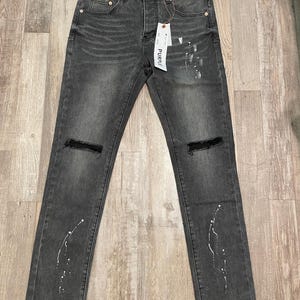 Op de afbeelding: Donkergrijze denim jeans met distressed kniedetails en witte verfspataccenten. De jeans hebben een klassiek ontwerp met vijf zakken en een knoop- en ritssluiting. Een label met het woord "PURPLE" is bevestigd.