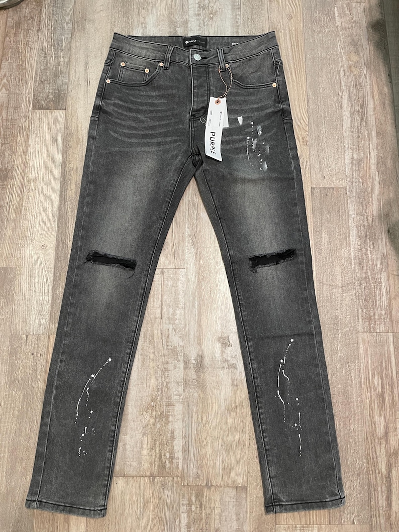 Op de afbeelding: Donkergrijze denim jeans met distressed kniedetails en witte verfspataccenten. De jeans hebben een klassiek ontwerp met vijf zakken en een knoop- en ritssluiting. Een label met het woord "PURPLE" is bevestigd.