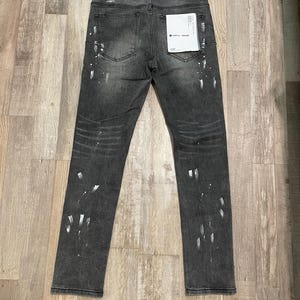 Op de afbeelding: Donkergrijze denim jeans met witte verfspatten. De jeans hebben een klassiek ontwerp met vijf zakken en een rechte pijp. Een klein label is bevestigd aan de achterzak. De jeans liggen plat op een houten oppervlak.